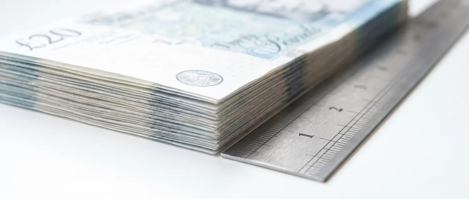 Stacked pale banknotes seen edge-on beside a metal ruler
